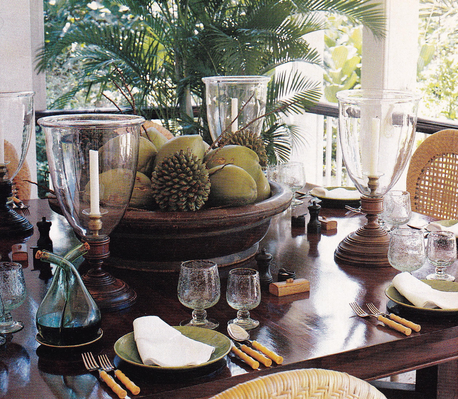 Dining table on the upper veranda with glass hurricane lamps, green fruit and woven chairs beside palms