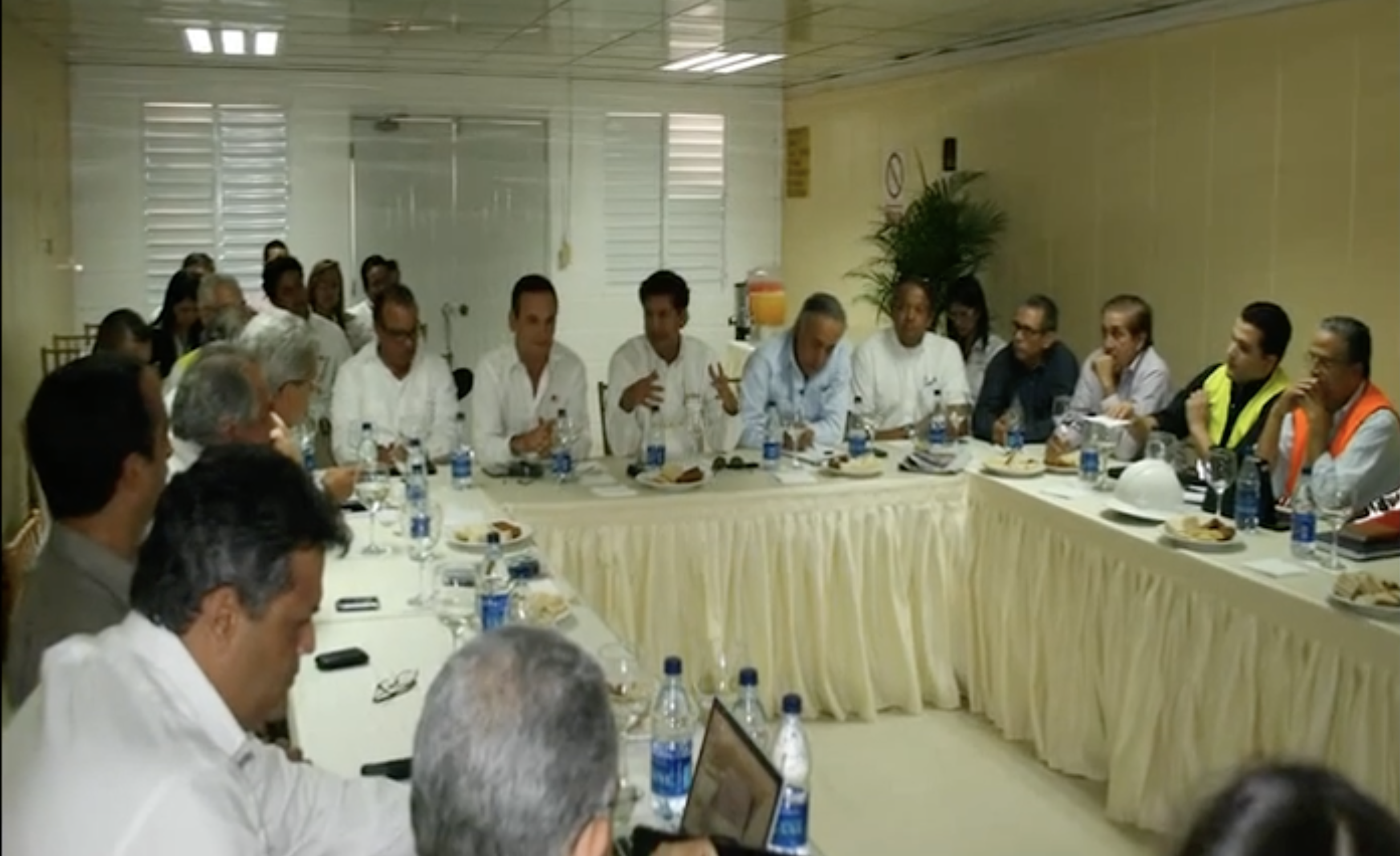 Meeting attendees seated around white-covered tables in the Cemento Panam documentary; Miguel H. Peña identifies himself and his father among those present